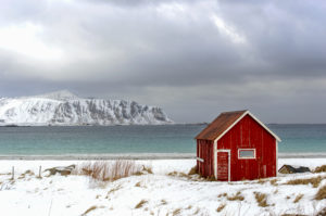 un rorbu rouge (chalet de pêcheur en bois) isolé au cœur d’un décor enneigé. Au loin, vue sur la mer et le fjord